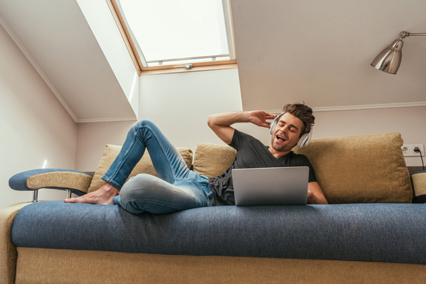 excited man listening music in wireless headphones while lying on sofa near laptop - Fotó, kép