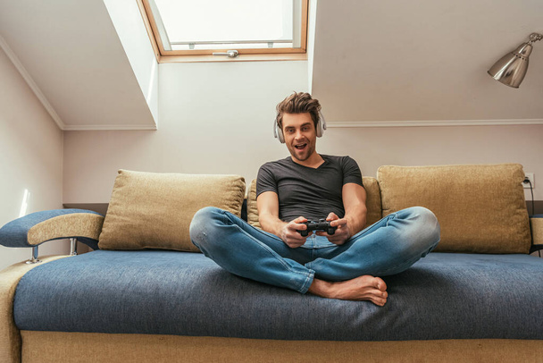 KYIV, UKRAINE - APRIL 13, 2019: excited man in wireless headphones on neck playing video game while sitting on sofa with crossed legs - Фото, изображение
