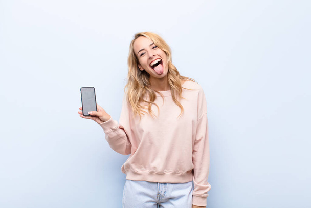 young blonde woman with cheerful, carefree, rebellious attitude, joking and sticking tongue out, having fun using a smartphone - Zdjęcie, obraz
