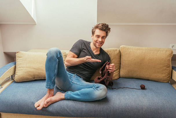 felice giovane uomo che punta con mano mentre lavora a maglia sul divano di casa
 - Foto, immagini