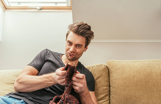 uomo scontento guardando la fotocamera mentre lavorava a maglia sul divano a casa
 - Foto, immagini
