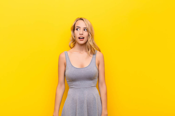 young blonde woman feeling shocked, happy, amazed and surprised, looking to the side with open mouth against orange wall - Foto, afbeelding