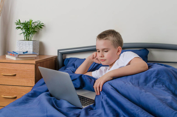 Дистанционное обучение и концепция онлайн образования. Школьник учится дома. Мальчик с ноутбуком лежит в постели. Социальное расстояние во время карантина
. - Фото, изображение