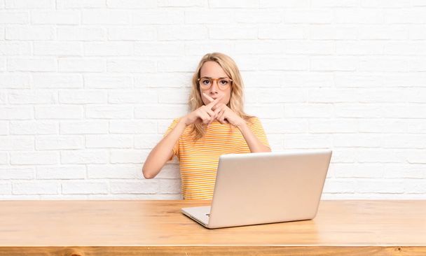 young blonde woman looking serious and displeased with both fingers crossed up front in rejection, asking for silence using a laptop - Fotoğraf, Görsel