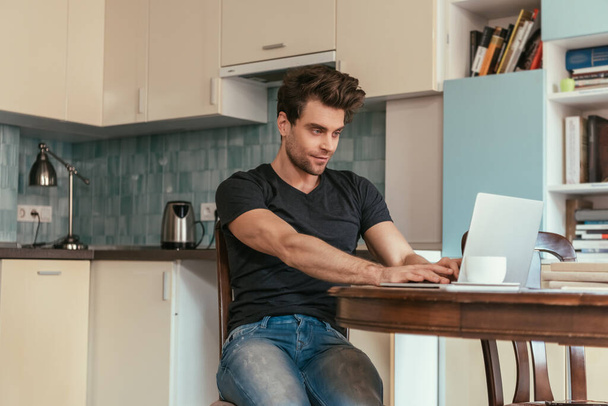 attentive, smiling man typing on laptop in kitchen - Foto, Imagen