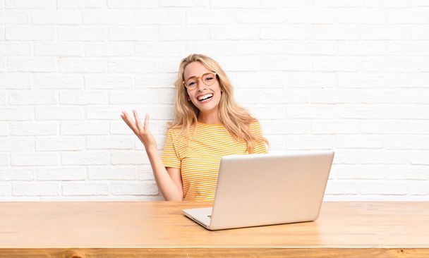 young blonde woman feeling happy, surprised and cheerful, smiling with positive attitude, realizing a solution or idea using a laptop - Foto, Imagen