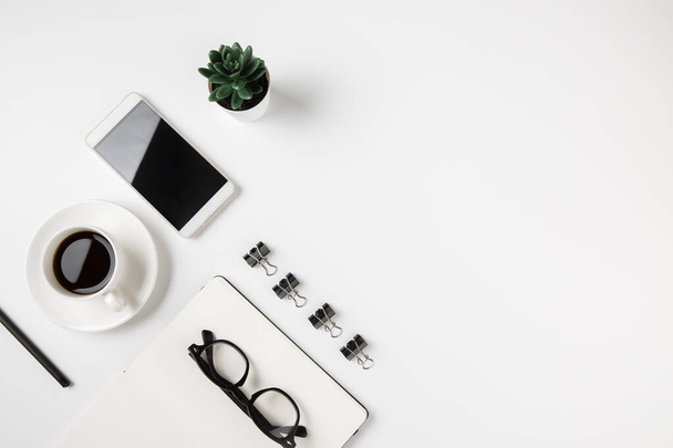 Λευκό minimal επιφάνεια εργασίας τραπέζι με ανοιχτή κενή σελίδα σημειωματάριο, φλιτζάνι καφέ, γυαλιά ματιών, smartphone, πράσινο χυμώδες φυτό. Αντιγραφή χώρου. - Φωτογραφία, εικόνα