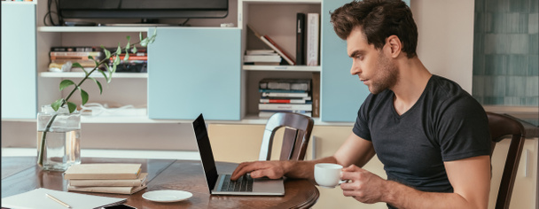 side view of concentrated man holding cup of coffee while working on laptop at home, panoramic shot - Photo, Image