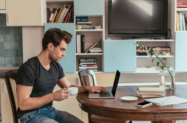 side view of thoughtful man holding cup of coffee while working at laptop in kitchen - Photo, Image
