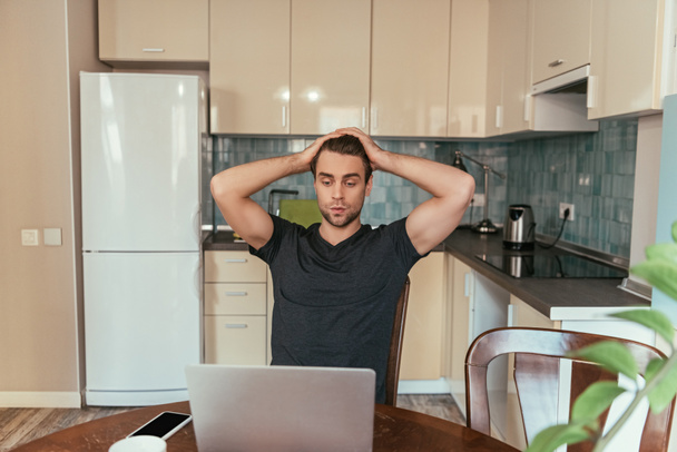 exhausted man touching head while looking at laptop monitor in kitchen - Foto, Imagen