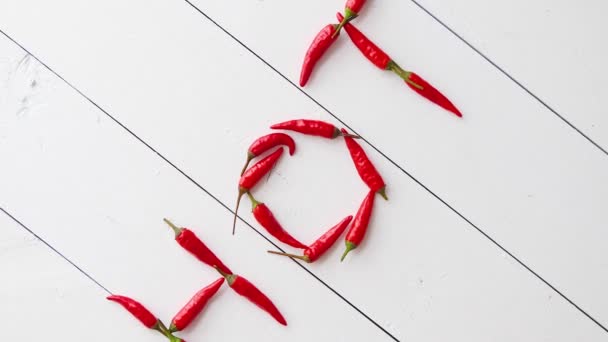 Slovo HOT vytvořené malými červenými chilli papričkami. Umístěno na bílém dřevěném stole - Záběry, video