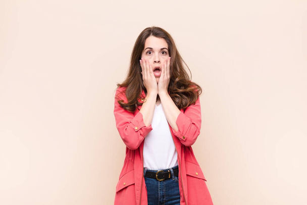 young pretty woman feeling shocked and scared, looking terrified with open mouth and hands on cheeks against beige background - Фото, изображение