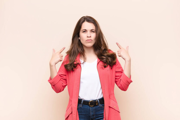 young pretty woman with a bad attitude looking proud and aggressive, pointing upwards or making fun sign with hands against beige background - Foto, imagen