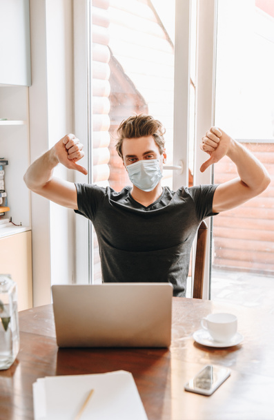 young man in medical mask showing thumbs down near laptop and cup of coffee on table - Photo, Image