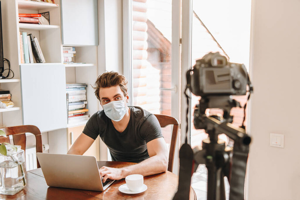 selective focus of young vlogger in medical mask looking at digital camera while sitting near laptop - Photo, Image