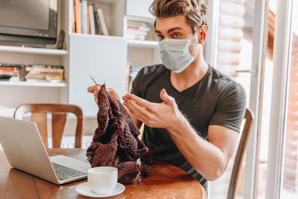 jovem em máscara médica sentado com os braços abertos perto de laptop e xícara de café enquanto segurando fios de tricô
 - Foto, Imagem
