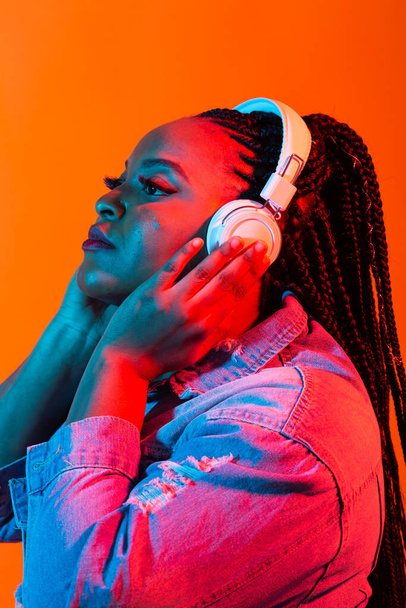 Close-up beauty fashion portret van een aantrekkelijke jonge Afrikaanse vrouw met dreadlocks op oranje achtergrond, luisterend naar muziek met hoofdtelefoon. Neonlicht. - Foto, afbeelding
