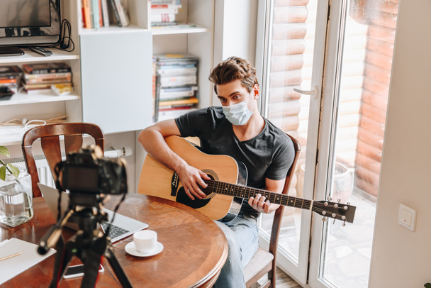 joven video blogger en máscara médica tocando la guitarra mientras mira la cámara digital en el trípode
 - Foto, Imagen