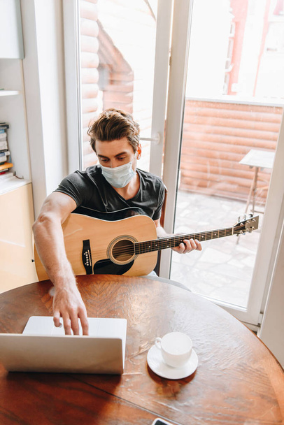 visão de alto ângulo do homem em máscara médica segurando guitarra e usando laptop em casa
 - Foto, Imagem