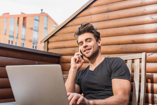 jóképű férfi használja laptop és beszél okostelefonon, miközben ül az erkélyen, és mosolyog a kamerába - Fotó, kép