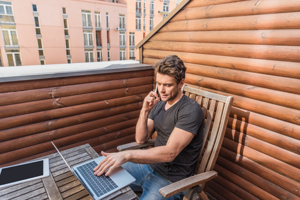 bonito, homem sério usando laptop e falando no smartphone enquanto sentado na varanda
 - Foto, Imagem