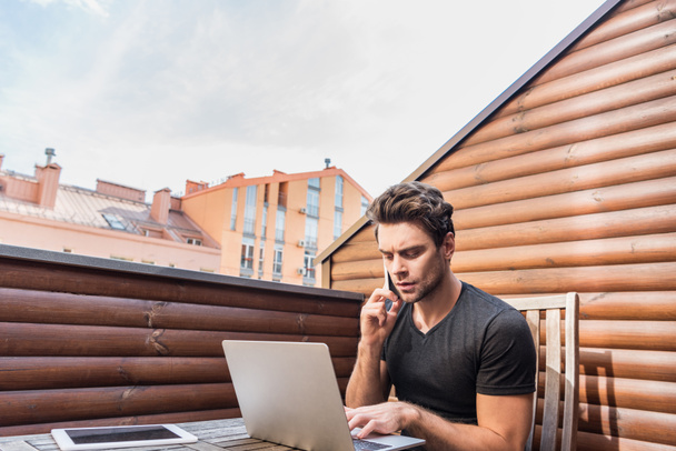 młody, poważny mężczyzna korzystający z laptopa i rozmawiający na smartfonie siedząc na balkonie - Zdjęcie, obraz