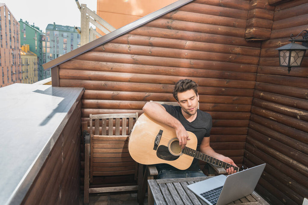 junger Mann mit Gitarre und Blick auf Laptop auf dem Balkon - Foto, Bild