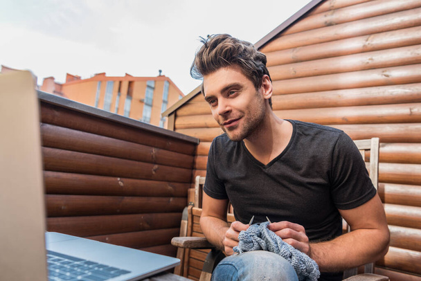 внимательный молодой человек смотрит на ноутбук во время вязания на балконе
 - Фото, изображение