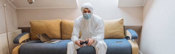 KYIV, UKRAINE - APRIL 13, 2019: panoramic shot of young man in hazmat suit and protective mask sitting on sofa near laptop and looking at camera - Foto, immagini