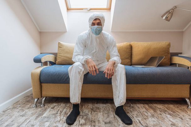 young man in hazmat suit and protective mask sitting on sofa near laptop and looking at camera - 写真・画像