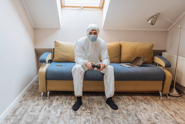 KYIV, UKRAINE - APRIL 13, 2019: young man in hazmat suit and protective mask playing video game while sitting on sofa near laptop and smartphone - Foto, imagen