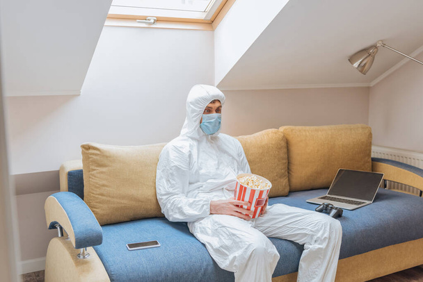 KYIV, UKRAINE - APRIL 13, 2019: man in hazmat suit and protective mask holding popcorn bucket while sitting on sofa near laptop, joystick and smartphone - Foto, afbeelding