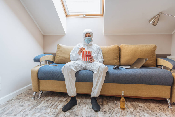 KYIV, UKRAINE - APRIL 13, 2019: young man in hazmat suit and protective mask sitting on sofa and eating popcorn near joystick and laptop - Zdjęcie, obraz