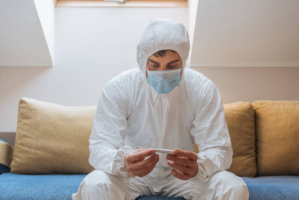 young man in hazmat suit and protective mask sitting on sofa and looking at thermometer - Photo, Image