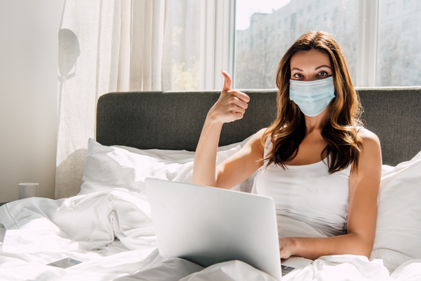 young freelancer in medical mask showing thumb up while working on laptop during self isolation in bed - Foto, Imagen