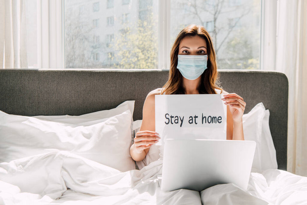 female freelancer in medical mask holding Stay at home sign and working on laptop during self isolation in bed - Photo, Image