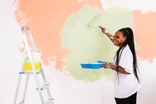 Joven mujer afroamericana feliz pintando la pared interior con rodillo de pintura en casa nueva. Una mujer con rodillo aplicando pintura en una pared. - Foto, imagen