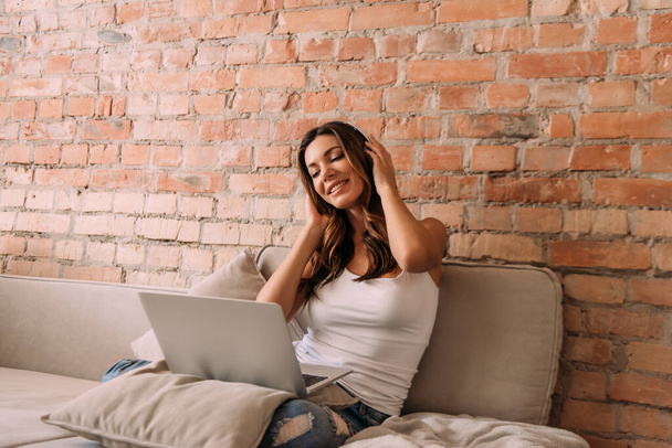 smiling girl listening music with headphones and laptop on sofa during self isolation - Foto, Bild