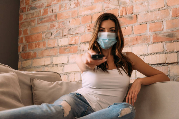 menina em máscara médica segurando controlador remoto e assistindo tv no sofá durante a quarentena, foco seletivo
 - Foto, Imagem