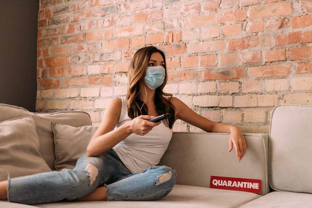 attractive girl in medical mask holding remote controller and watching tv on sofa with quarantine sign  - Zdjęcie, obraz