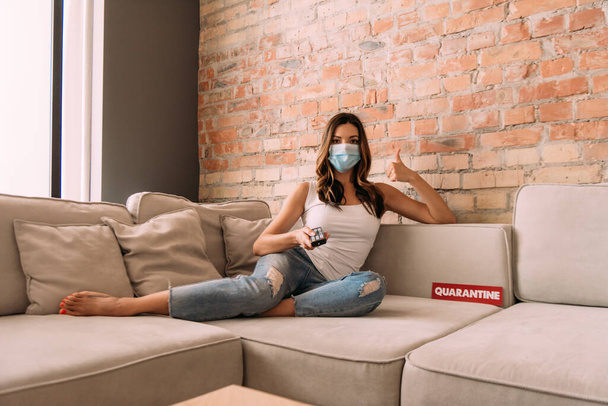 woman in medical mask showing thumb up and holding remote controller while watching tv on sofa with quarantine sign  - Photo, image