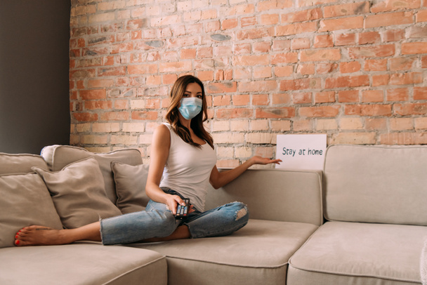 woman in medical mask holding remote controller and watching tv on sofa with stay at home sign during self isolation - Valokuva, kuva
