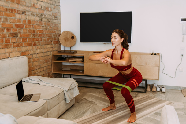 beautiful sportswoman squating with resistance band on fitness mat, training online with laptop during self isolation  - Photo, Image