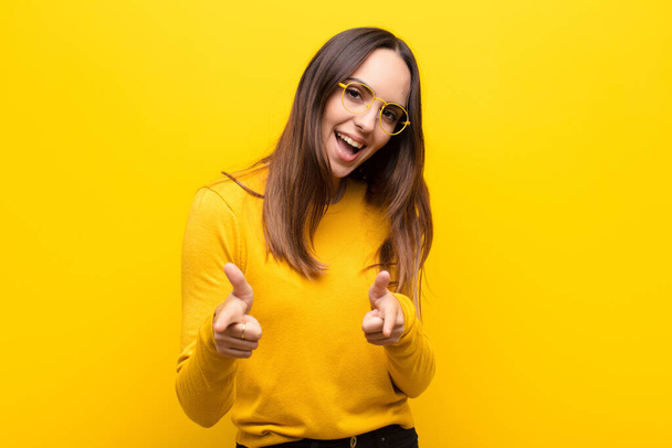 young pretty woman feeling happy, cool, satisfied, relaxed and successful, pointing at camera, choosing you against orange wall - Фото, изображение