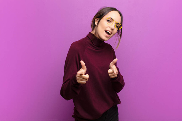 young pretty woman feeling happy, cool, satisfied, relaxed and successful, pointing at camera, choosing you against purple wall - Foto, Bild
