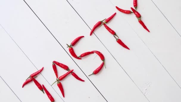 Słowo TASTY utworzone z małych czerwonych papryczek chili. Umieszczony na białym drewnianym stole - Materiał filmowy, wideo