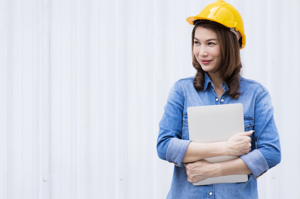 Beautiful Asian female engineer in jeans dress wearing yellow safety hard hat using laptop computer notebook doing job at construction site outside office. Idea for modern working woman. - Photo, Image