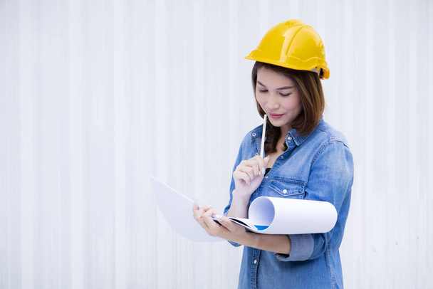 Beautiful Asian female engineer in jeans dress wearing yellow safety hard hat doing job at construction site outside office. Idea for modern working woman and outdoor work career of worker. - Photo, Image