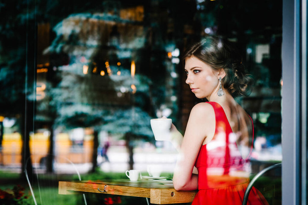 Beautiful serious European brunette girl in an evening red dress sitting at the window with a cup of coffee at the cafe. Looking at the side through the window. - Фото, изображение