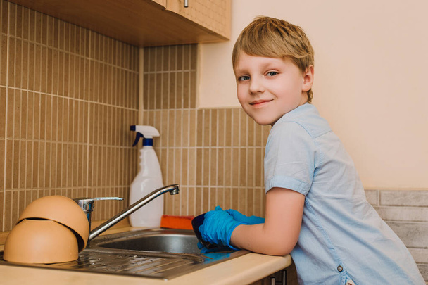 Хлопчик допомагає мамі виконувати домашню роботу. Хлопчик в захисних синіх гумових рукавичках миє чашки на кухні
. - Фото, зображення